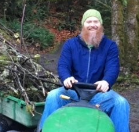 Picture of a bearded masculine person named Christopher, wearing a hat, smiling, and sitting on a small tractor in nature